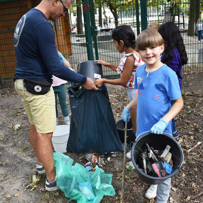 Bild vergrößern: Hort Wirbelwind - Herbstputz 2023 -  mehrere Kinder und ein Vater füllen den in Eimern gesammelten Müll in einen blauen Müllsack