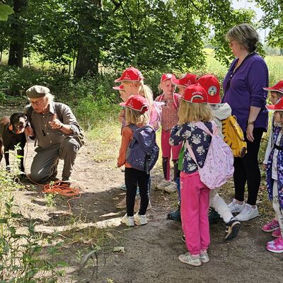 Bild vergr&ouml;&szlig;ern: Der J&auml;ger kniet neben seinem Jagdhund und die Kinder schauen zu.