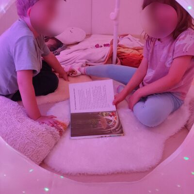 Bild vergr&ouml;&szlig;ern: Zwei Kinder sitzen gemeinsam in einer gem&uuml;tlichen wei&szlig;en H&ouml;hle auf weichen Lammfellen und Kissen. Sie schauen sich gemeinsam ein Bilderbuch an. Das Licht ist beruhigend rosa-violett.