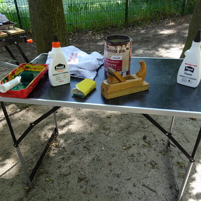 Bild vergr&ouml;&szlig;ern: Auf einem Tisch befindet sich ein Teil der verwendeten Materialien (z. B. Holzleim, Wetterschutzholzfarbe, Hobel, Schleifpapier, Farbrolle).