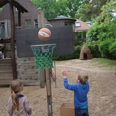 Bild vergr&ouml;&szlig;ern: Ein Kind mit einem langen Zopf sowie ein Kind mit blauer Jacke stehen zum Basketballkorb und der Ball fliegt gerade in den Korb hinein.