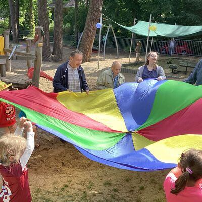 Bild vergr&ouml;&szlig;ern: Erwachsene und Kinder stehen lachend an einem bunten Schwungtuch und halten dieses nach oben.