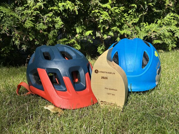 Stadtradeln-Siegerpokal 2026 vor einem dunkelblau-roten und einem mittelblaufarbenem Fahrradhelm auf einer Wiese.