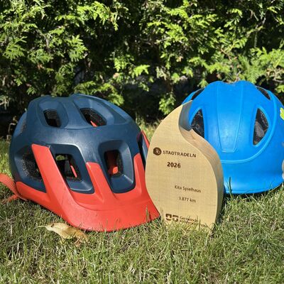 Stadtradeln-Siegerpokal 2026 vor einem dunkelblau-roten und einem mittelblaufarbenem Fahrradhelm auf einer Wiese.